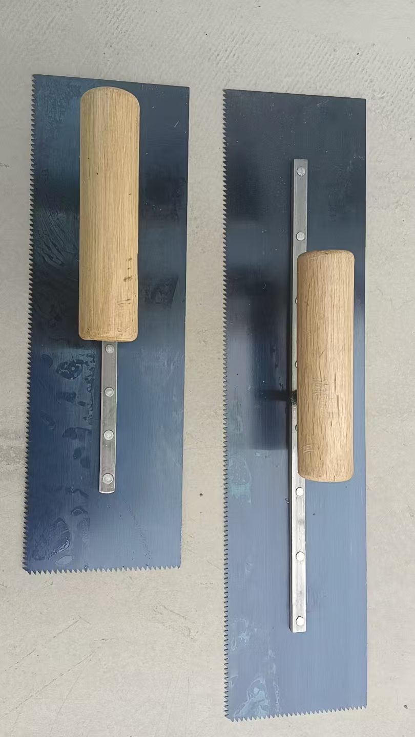 Two blued steel notched trowels with wooden handles, laid on a concrete slab